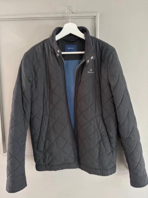 Svart quiltad jacka från Gant - Stilren svart quiltad jacka från Gant med ståkrage och dragkedja framtill. Jackan har diskret logga på bröstet och ett klassiskt rutmönster. Perfekt för dig som vill ha en snygg och enkel jacka till vardags.