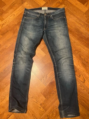 Blå raka jeans från Acne Studios - Säljer ett par blå jeans från Acne Studios med raka ben och klassisk femficksmodell. Jeansen har snygga slitningar och tvättade detaljer för en avslappnad look. Tillverkade i denim med kontrastsömmar och knappgylf.