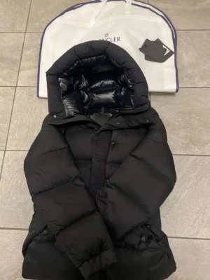 Svart dunjacka från Moncler - Snygg svart dunjacka från Moncler med stor, vadderad huva och diskreta sidofickor. Jackan har en glansig finish och stängs med både dragkedja och knappar. Perfekt för kalla vinterdagar och riktigt varm tack vare dunfyllningen.