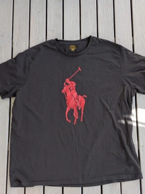 Svart t-shirt med röd polospelare. Big Pony. 3XL - Snygg svart t-shirt med en stor röd polospelare tryckt på bröstet. Klassisk rund hals och korta ärmar. Tillverkad i mjuk bomull som känns skön mot huden. Perfekt för dig som gillar stilrena plagg med en sportig touch. Model 515219