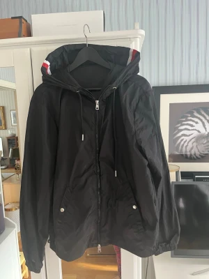 Svart vindjacka från Moncler - Snygg svart vindjacka från Moncler med huva och dragkedja framtill. Jackan har två fickor med tryckknappar och vita/röda detaljer på huvan. Moncler-logga på ärmen. Perfekt för blåsiga dagar och enkel att matcha.