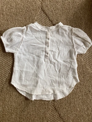 Vit blus med puffärm från Zara - Säljer en vit blus från Zara i mjukt, krinklat bomullstyg. Blusen har puffiga korta ärmar, rundad halsringning och knappar framtill. Perfekt till jeans eller kjol för en avslappnad men söt look.