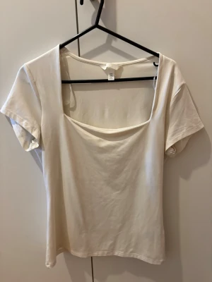 Vit topp med fyrkantig ringning - Stilren vit/beige topp med korta ärmar och fyrkantig ringning. Tillverkad i ett mjukt och lätt material som känns skönt mot huden. Perfekt att matcha med jeans eller kjol för en enkel och snygg look.
