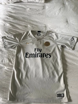 PSG Mbappé matchtröja vit - Snygg vit PSG matchtröja från Nike med guldiga och svarta detaljer. Framsidan har Fly Emirates-tryck och PSG-logga, baksidan har Mbappé och nummer 7. Korta ärmar och sportig passform, perfekt för fotboll eller träning. Pris kan diskuteras.