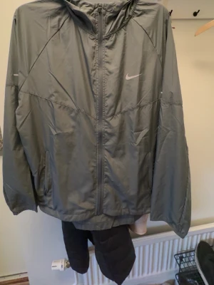 Grå vindjacka med huva från Nike Nike Miller - Snygg grå vindjacka från Nike med dragkedja framtill och praktisk huva. Jackan har två sidofickor och diskret Nike-logga på bröstet. Tillverkad i lätt och vindtätt material, perfekt för blåsiga dagar.