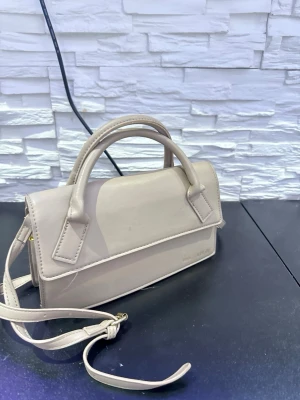 Beige minihandväska med axelrem - Snygg beige minihandväska i slätt konstläder med både handtag och justerbar axelrem. Väskan har en stilren, rektangulär form och guldfärgade detaljer. Perfekt för dig som gillar minimalistisk och trendig design.
