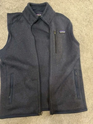 Mörkgrå fleeceväst från Patagonia - Mysig mörkgrå fleeceväst från Patagonia med hög krage och dragkedja framtill. Västen har tre fickor med dragkedja och en stilren look som passar perfekt till lager-på-lager. Perfekt för kyliga dagar.