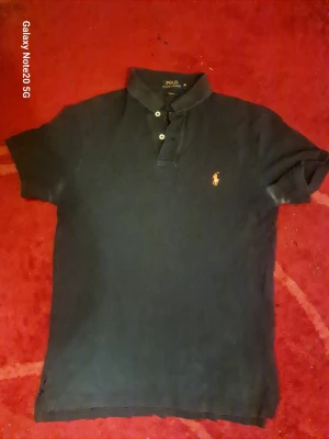 Svart pikétröja från Polo Ralph Lauren - Klassisk svart pikétröja från Polo Ralph Lauren med korta ärmar och slim fit-passform. Tröjan har krage, knappar framtill och den ikoniska broderade loggan på bröstet. Perfekt till jeans eller shorts för en clean och stilren look.