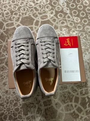 Christian Louboutin - Snygga gråa sneakers i mocka från Christian Louboutin med vita sulor och klassisk låg profil. Skorna har matchande grå skosnören och diskreta sömdetaljer. Insidan är fodrad i beige läder och märket syns på innersulan. Perfekta för en stilren och modern look.