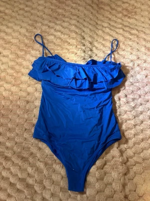 Blå baddräkt med volang - Snygg blå baddräkt med smala axelband och volangdetalj över bysten. Klassisk modell med en feminin touch, perfekt för stranden eller poolen. Materialet är stretchigt och bekvämt för en bra passform.