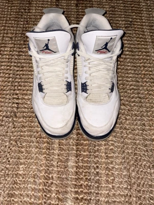 Vita Air Jordan 4 sneakers - Säljer ett par vita Air Jordan 4 sneakers med marinblå detaljer och meshpaneler på sidorna. Skorna har klassisk Jumpman-logga på plösen och en robust sula med synlig Air-enhet. Perfekta för dig som gillar retrostil och streetwear.