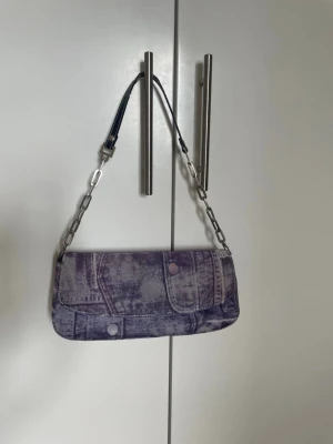 Lila denim-inspirerad handväska - Handväska från Jet Mode i lila denim-look med tryckt jeansmönster. Väskan har en silverfärgad kedjerem och insidan är beige med dragkedjefack. Hör av er vid frågor eller prisförslag! 
