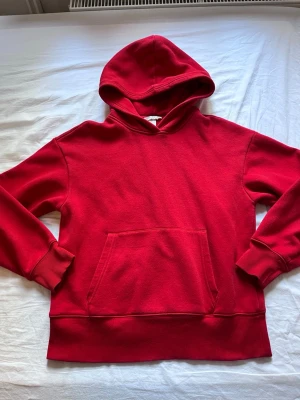 Röd hoodie från H&M med ficka - Säljer en klassisk röd hoodie från H&M i storlek S. Hoodien har en stor känguruficka framtill, ribbade muddar vid ärmar och nederkant samt en skön huva. Perfekt för dig som gillar enkel och stilren streetwear.