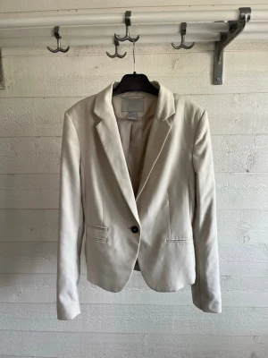 Beige kavaj från H&M, storlek 34 - Stilren beige kavaj från H&M i storlek 34. Klassisk modell med en knapp framtill, smala slag och två fickor. Tillverkad i ett mjukt tyg med slät finish och helfodrad insida. Perfekt för att lyfta din outfit med en clean look.