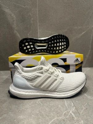 Vita Adidas Ultraboost sneakers - Säljer ett par fräscha vita Adidas Ultraboost sneakers med stickad ovandel och klassiska tre ränder på sidan. Skorna har en bekväm Boost-sula och svart yttersula. Perfekta för dig som gillar stilrena och sportiga sneakers.