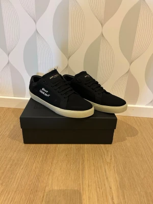 Svarta Saint Laurent sneakers - Snygga svarta sneakers från Saint Laurent med vit sula och diskret logga på sidan. Skorna har klassisk låg profil, svarta skosnören och ovandel i mocka. Perfekta för dig som gillar stilrena och tidlösa sneakers.