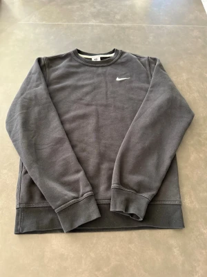 Svart sweatshirt från Nike - Klassisk svart sweatshirt från Nike med rund halsringning och diskret vitt Nike-logga broderad på bröstet. Tröjan har ribbade muddar vid ärmslut och nederkant, perfekt för en avslappnad och sportig stil.