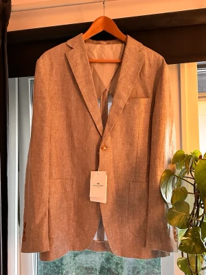 Beige linnékavaj från John Henric - Säljer en stilren beige kavaj från John Henric i storlek 50.  Kavajen är helt ny och har kvar originaltaggen, vilket innebär att du får en kavaj i nyskick utan att behöva betala fullpris i butik.  - Nypris: 2 000 kr  Kavajen är tillverkad i en exklusiv linne- och bomullsblandning och passar perfekt under vår och sommar. Den klassiska beige färgen gör den väldigt lätt att matcha – lika snygg till kostymbyxor och skjorta som till jeans och en mer avslappnad outfit.   • Storlek: 50 EU