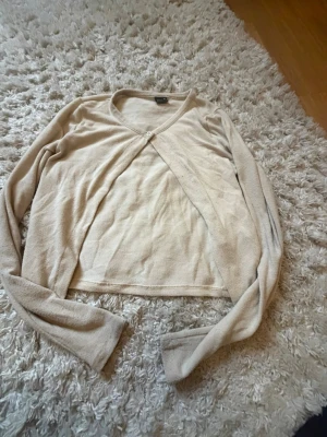 Beige tunn kofta med knapp - Ljusbeige tunn kofta med v-ringning och en enkel knapp framtill. Modellen är croppad med långa ärmar och mjukt, lätt material. Perfekt att slänga över ett linne eller t-shirt för en avslappnad look.