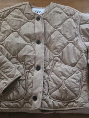 Beige quiltad cardigan från ZARA - Snygg beige quiltad cardiganjacka från ZARA i storlek XS. Jackan har rund halsringning, stora framfickor och stängs med svarta knappar. Materialet är mjukt och lätt vadderat, perfekt för lager på lager. Stilsäker och trendig modell med rutmönstrad quiltning.