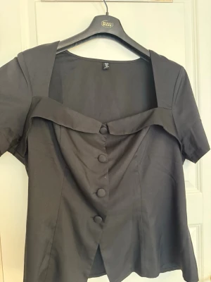 Svart blus med stora knappar - Stilren svart blus med korta ärmar och fyrkantig urringning. Framtill har den stora klädda knappar och en dekorativ krage som ger en unik look. Materialet ser ut att vara mjukt och lätt, perfekt för en trendig outfit.