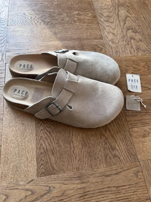 Beige mockatofflor från Pace - Säljer ett par snygga beige tofflor från Pace i mjuk mocka. De har en stilren design med rund tå, justerbart spänne i metall och öppen häl. Perfekta för dig som gillar minimalistisk stil och vill ha något bekvämt att glida runt i.