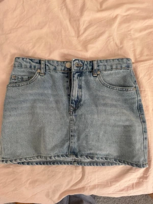 Ljusblå jeanskjol med fickor - Säljer en klassisk ljusblå jeanskjol i kort modell. Kjolen har fem fickor, bälteshällor och stängs med knapp och dragkedja framtill. Perfekt till sommaren och enkel att matcha med olika toppar.