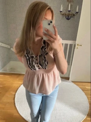 Ljusrosa blus med volang och knappar - Söt ljusrosa blus med volangdetaljer och knappar framtill. Blusen har smock i midjan som ger en fin form och korta ärmar. Perfekt för dig som gillar romantiska detaljer och en feminin look.