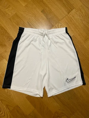 Vita shorts från Nike - Säljer dessa snygga vita sportshorts från Nike med svarta sidopaneler och klassisk Nike-logga på vänster ben. Shortsen har elastisk midja med snörning för bästa passform och är tillverkade i ett lätt och svalt syntetmaterial, perfekta för träning eller sport. Vid frågor, extra bilder eller liknande är det bara att kontakta mig!