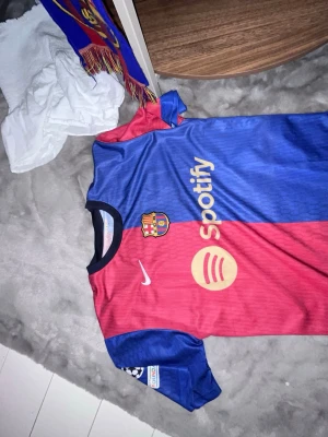 Barcelona matchtröja Lamine Yamal - Säljer en officiell FC Barcelona matchtröja i rött och blått med Lamine Yamal och nummer 27 på ryggen. Tröjan har Spotify-logga på bröstet, UNHCR ACNUR-tryck bak och Champions League-märke på ärmen. Tillverkad i lätt och ventilerande material. Inte en äkta tröja!