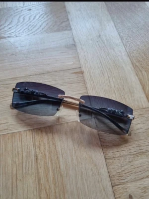 Svarta solglasögon med gulddetaljer - Snygga solglasögon med rektangulära, svarta glas och guldfärgade bågar. På sidorna finns en cool panterdetalj i guld som ger en unik och lyxig känsla. Glasen är tonade och bågarna är tunna och eleganta. Perfekt accessoar för att höja din stil.