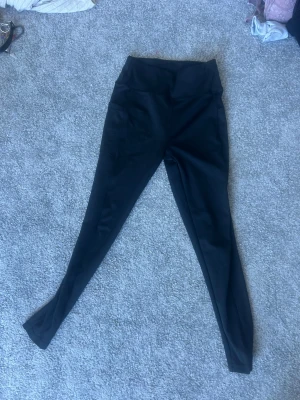 Svarta leggings med hög midja - Snygga svarta leggings med hög midja och praktisk ficka på sidan. Tillverkade i stretchigt material som sitter skönt och formar sig efter kroppen. Perfekta för både träning och chill. Passar dig som gillar enkel och stilren look.