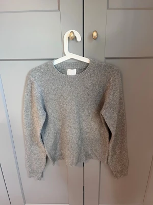 Grå stickad tröja från H&M - Mysig grå stickad tröja från H&M med rund halsringning och ribbade muddar vid ärmslut och nederkant. Perfekt för lager på lager och enkel att matcha med olika outfits. Klassisk modell i mjukt material. Lite nopprig 
