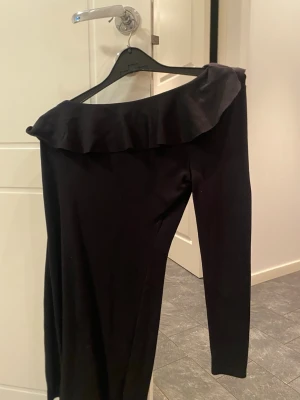 Svart oneshoulder klänning  - Säljer en svart oneshoulder klänning från Divided med volangdetalj längs urringningen. Klänningen har lång ärm på ena sidan och är axelbandslös på andra. Perfekt för dig som vill ha en stilren och trendig look.