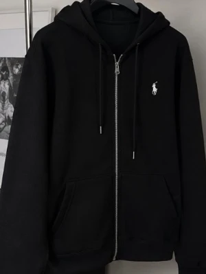 Svart zip hoodie med logga - Snygg svart hoodie med dragkedja och huva. Har en vit broderad logga på bröstet och praktiska fickor framtill. Tillverkad i mjukt material, perfekt för en avslappnad stil. Dragsko i huvan för justerbar passform.