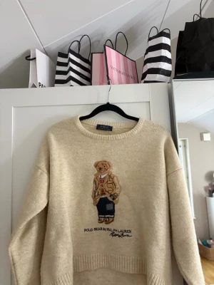 Stickad Polo Ralph Lauren Bear tröja  - Stickad beige tröja från Polo Ralph Lauren med den ikoniska Polo Bear-broderingen framtill. Tröjan har rund halsringning, ribbade muddar och är gjord i mjukt material. 