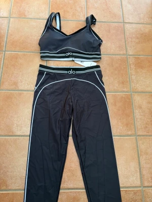 Alo Yoga Set - Snyggt svart set från Alo med sport-bh och matchande tights. Setet har vita kontrastsömmar och logga på både bh och midjeband. Materialet är stretchigt och följsamt, perfekt för en aktiv livsstil. Modern design med sportig känsla.
