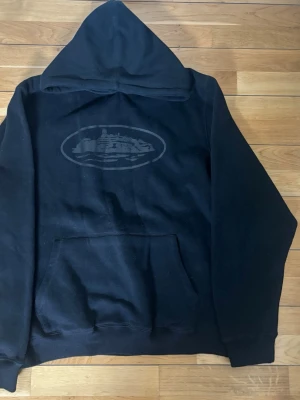 Svart hoodie med tryck framtill från Cortiez - Svart hoodie med stor huva och magficka. På bröstet finns ett diskret tryck med motiv av en byggnad inuti en oval. Hoodien har ribbade muddar vid ärmar och nederkant. Perfekt för en avslappnad och cool stil.