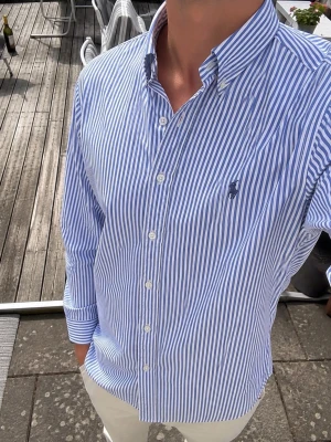Ralph Lauren Classic Fit – Blå/Vit Randig Skjorta - Stilren Ralph Lauren Classic Fit skjorta i klassiskt blå- och vitrandigt mönster. Med ikonisk broderad hästlogga på bröstet och button-down-krage som ger en tidlös, preppy look. Perfekt att bära till chinos, jeans eller shorts för en klassisk old money-inspirerad stil. Skjortan är endast använd en gång och är därför i mycket fint skick, i princip som ny. Storlek M.