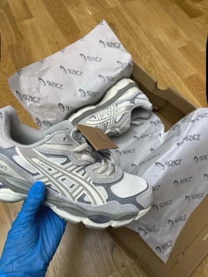 Gråvita sneakers från Asics - Snygga sneakers från Asics i grått och vitt med mesh- och syntetpaneler. Skorna har en chunky sula och klassisk Asics-logga på sidan. Perfekta för dig som gillar sportig och trendig stil.