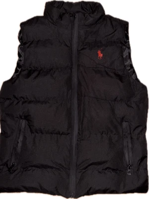 Svart dunväst med röd logga - Snygg svart dunväst från US Polo Assn med hög krage och dragkedja framtill. Västen har två fickor med dragkedja och en röd broderad logga på bröstet. Perfekt för lager på lager och streetwear-stil. Skriv ifall ni har funderingar. OBS fick denna från min kusin så vet inte om den är äkta 