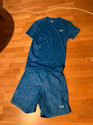 Blått träningsset från Under Armour - Säljer ett blått träningsset från Under Armour med t-shirt och shorts. T-shirten är enfärgad med logga på bröstet och shortsen har ett diskret mönster samt logga på benet. Perfekt för gymmet eller löpning.