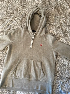 Grå Ralph Lauren hoodie med röd logga - Snygg grå hoodie från Polo Ralph Lauren med klassisk röd broderad logga på bröstet. Hoodien har huva med vita snören och en stor magficka framtill. Tillverkad i mjukt sweatshirtmaterial.