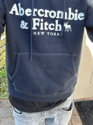 Mörkblå hoodie från Abercrombie & Fitch - Snygg mörkblå hoodie från Abercrombie & Fitch med stor vit logga och texten 'New York' på bröstet. Klassisk känguruficka framtill och justerbar huva med snören. Perfekt för en avslappnad och cool stil. OM DU ÄR MELLAN 170cm och 172  kommer den passa perfekt