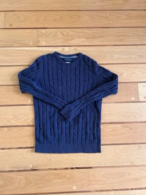 Mörkblå kabelstickad tröja Jack & Jones - Säljer en mörkblå kabelstickad tröja från Jack & Jones. Tröjan har rund halsringning, ribbade muddar och ett klassiskt flätmönster över hela plagget. Perfekt för dig som gillar stilrena och tidlösa plagg i garderoben.