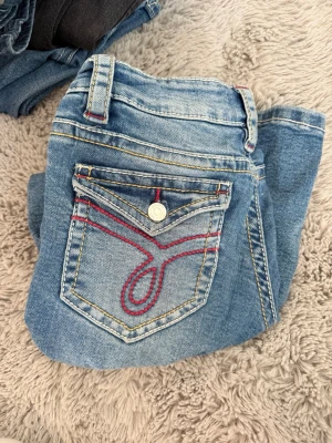 Blå jeans med röd söm och fickdetalj - Snygga ljusblå jeans med coola röda sömmar och unik fickdetalj bak. Klassisk femficksmodell med dragkedja och knapp. Jeansen har slitningar framtill för en trendig look och kontrastsömmar som sticker ut. Från Gina i storlek 158.