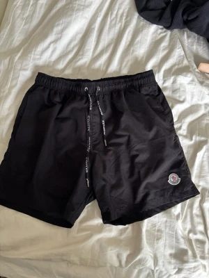 Svarta badbyxor med Moncler-logga - Snygga svarta badbyxor från Moncler med elastisk midja och dragsko med logotypdetaljer. Klassisk passform, Moncler-märke på vänster ben och en bakficka med dragkedja och rödvit detalj. Perfekta för stranden eller poolen.