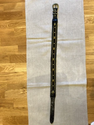 Svart bälte med nitar och mönster 92cm - Snyggt svart bälte i skinn med guldfärgade nitar längs hela bandet och präglat mönster på ändarna. Perfekt för att ge outfiten en edgy touch. Spännet är också i guldton och bältet har en smal form.