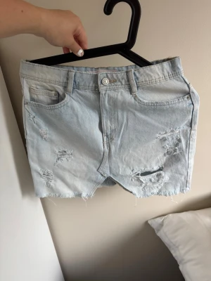 Ljusblåa slitna jeansshorts - Säljer ett par ljusblå jeansshorts med slitna detaljer och fransig kant nertill. Shortsen har klassisk femficksdesign och knappgylf. Perfekta för en avslappnad och trendig look i sommar.