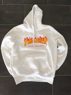 Vit Thrasher hoodie med flames - Står XL skulle säga att det passar L, eller S/M om man gillar väldigt oversized. Säljer en vit hoodie från Thrasher med den ikoniska flames-loggan i gult och rött på bröstet. Hoodien har stor huva, magficka och ribbade muddar. Perfekt för dig som gillar streetwear och skatekultur.